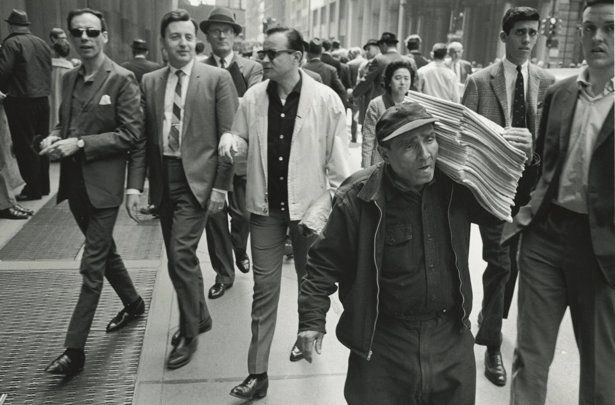 Black and white photograph of a group of people walking down a sidewalk. Man bottom right carrying a stack of newspapers.